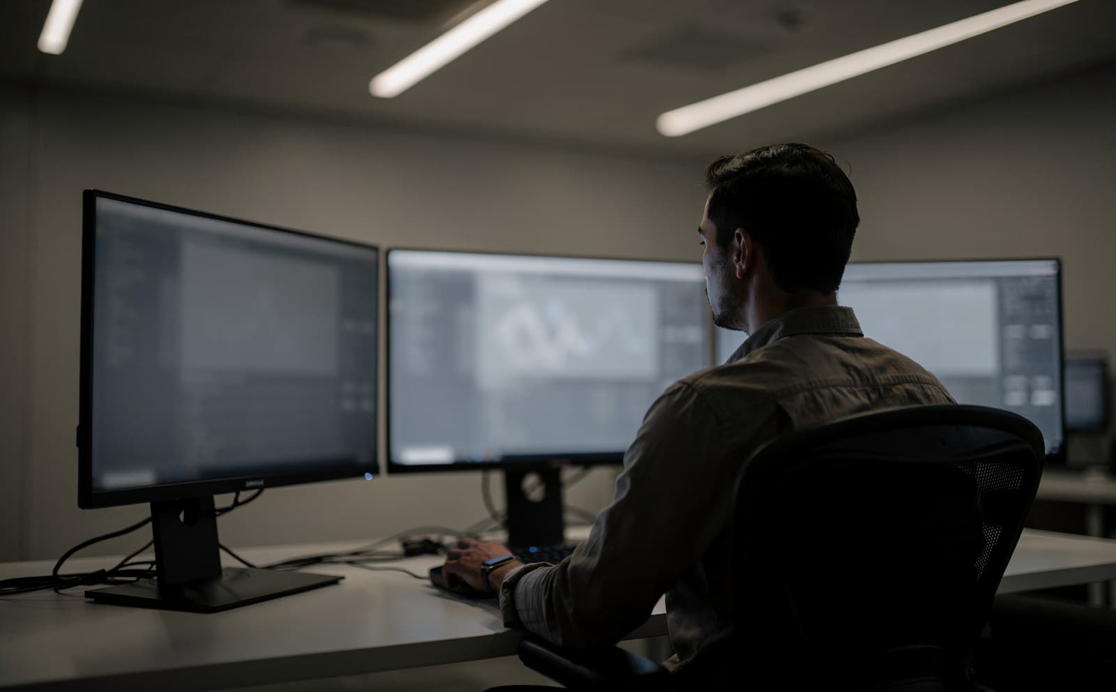 A calm modern GSOC operator working at a wide technical workstation with three understated monitors in soft ambient lighting