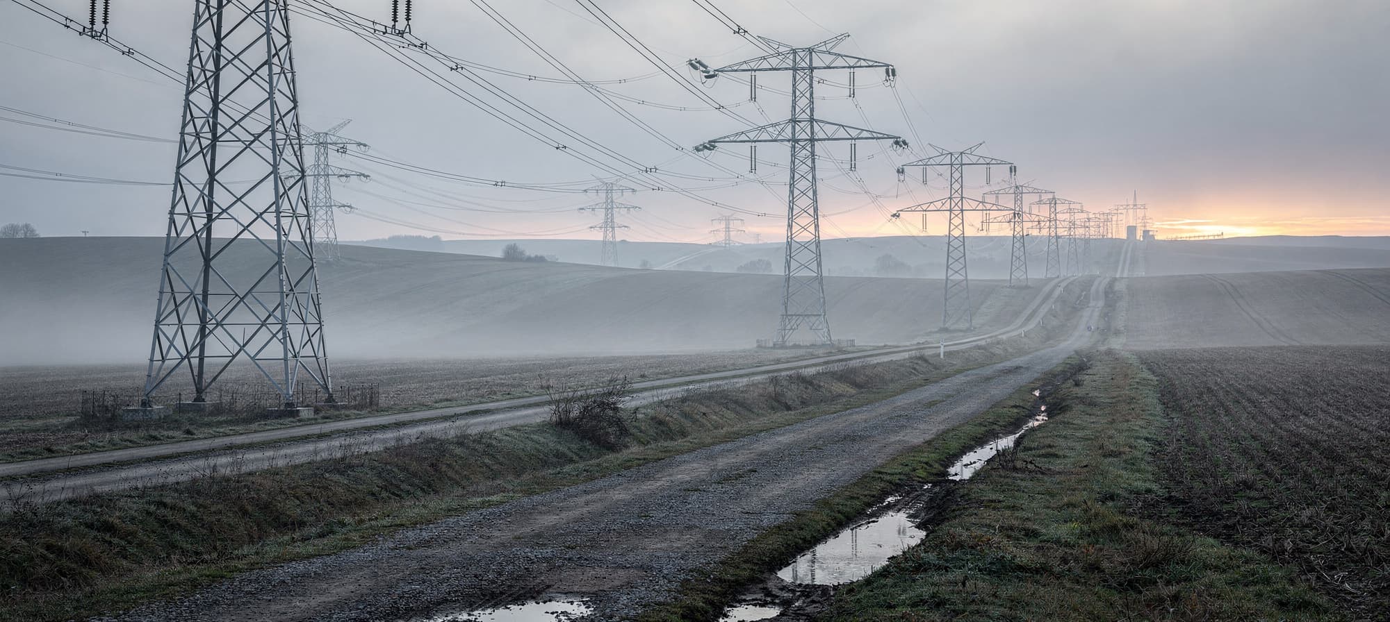 A wide high-voltage transmission corridor cutting across rolling farmland at first light, low ground fog, lattice steel towers marching into the distance